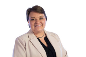A person with short brown hair is smiling, wearing a light beige blazer, a black top, and a pearl necklace, posing in front of a plain white background.