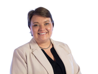 A person with short brown hair is smiling, wearing a light beige blazer, a black top, and a pearl necklace, posing in front of a plain white background.