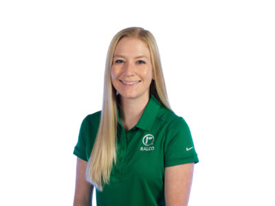A woman with long blond hair smiles at the camera. She is wearing a green polo shirt with the Ralco logo and a Nike logo on the sleeve, standing against a plain white background.