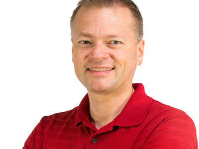 A smiling man with short hair wearing a red polo shirt stands with arms crossed against a plain white background.