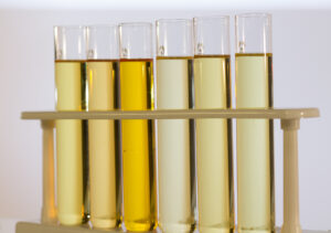 Six test tubes filled with liquids of varying yellow shades are held in a beige test tube rack against a plain white background.