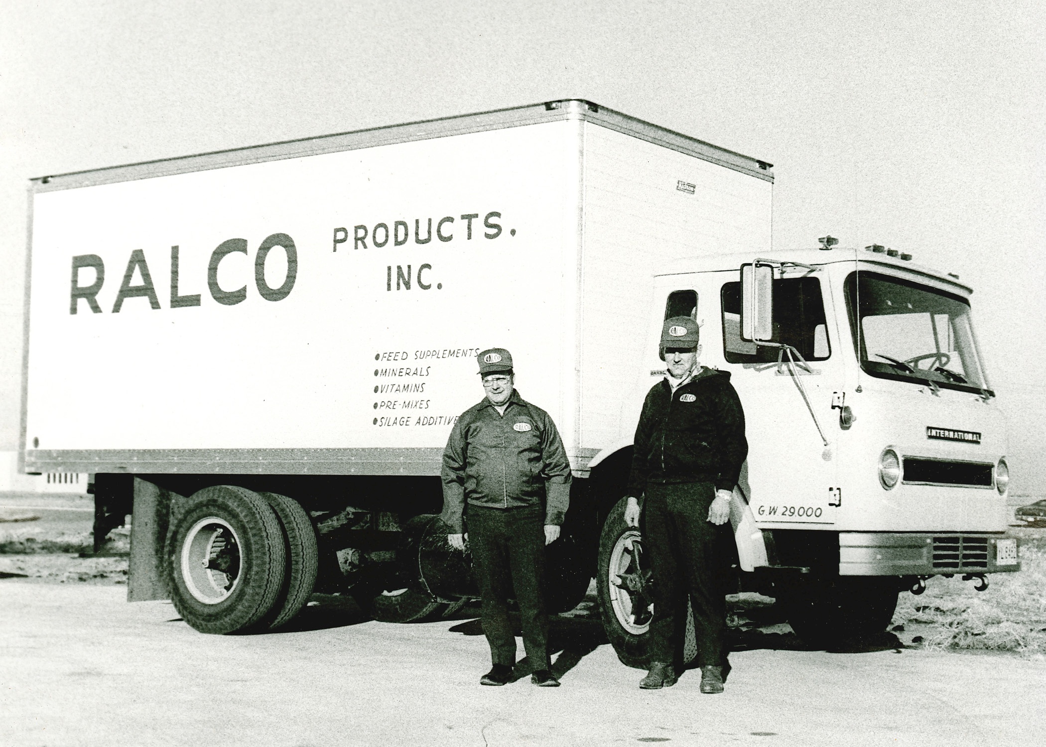 Two men in uniforms stand in front of a vintage box truck labeled "RALCO PRODUCTS, INC." The truck is white with large text and a list of products such as feed supplements and minerals on its side.