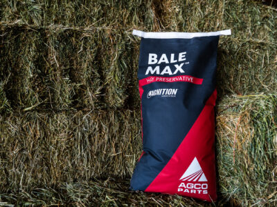 A bag labeled "Bale Max Hay Preservative" by AGCO Parts rests upright against stacked bales of hay. The bag is black and red with white text and logos.