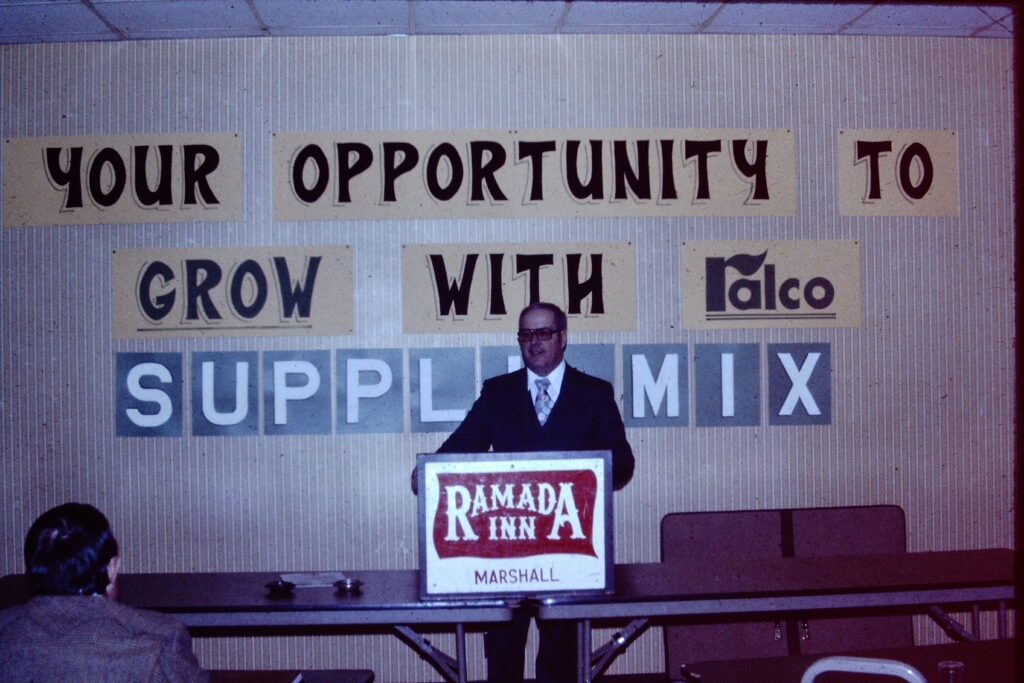 A man in a suit stands at a podium labeled "Ramada Inn Marshall," with a sign behind him reading "YOUR OPPORTUNITY TO GROW WITH Falco SUPPLEMIX." Two people are seated in front of the podium.