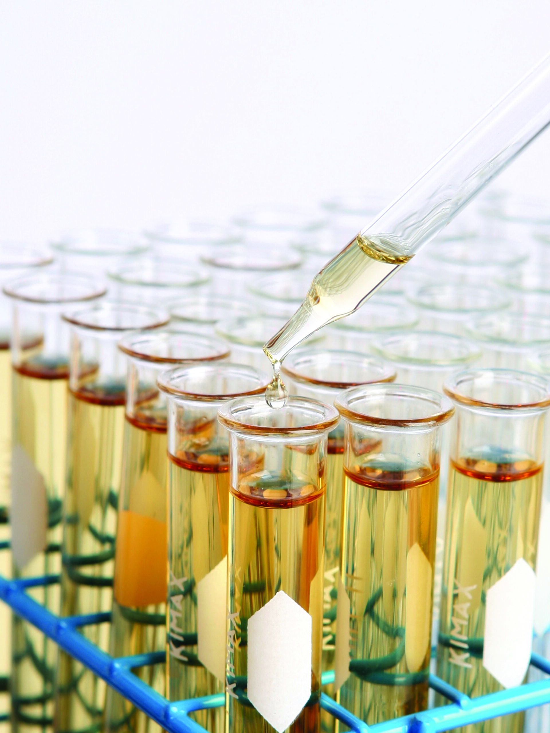A dropper releases liquid into one of several test tubes filled with a yellow solution, all held in a blue test tube rack in a laboratory setting.