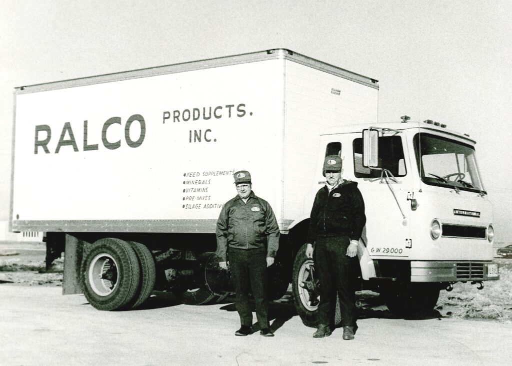 Two men in work uniforms stand in front of a large box truck labeled "RALCO PRODUCTS, INC." The truck is white with black text and parked on a paved surface, with both men smiling at the camera.