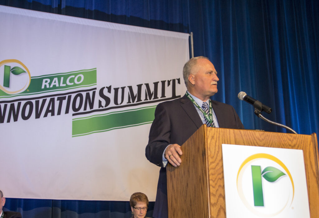 A man in a suit speaks at a podium with a microphone during the Ralco Innovation Summit, with a large event banner displayed behind him on a blue curtain background.