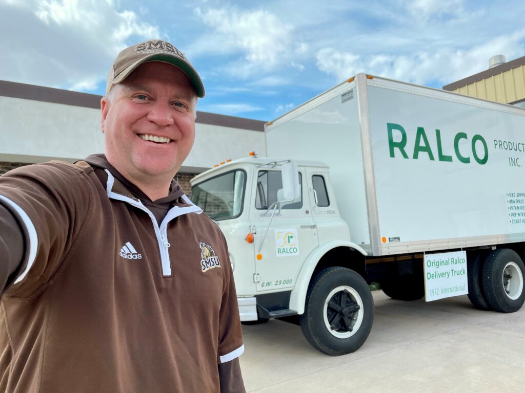 A man in a brown and white sports jacket and cap smiles for a selfie outside, standing near a white delivery truck labeled "RALCO." The truck is parked by a building under a partly cloudy sky.
