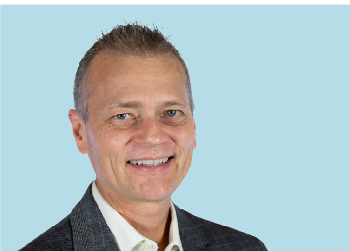 A middle-aged man with short, light brown hair wearing a gray checked blazer and white shirt smiles against a light blue background.