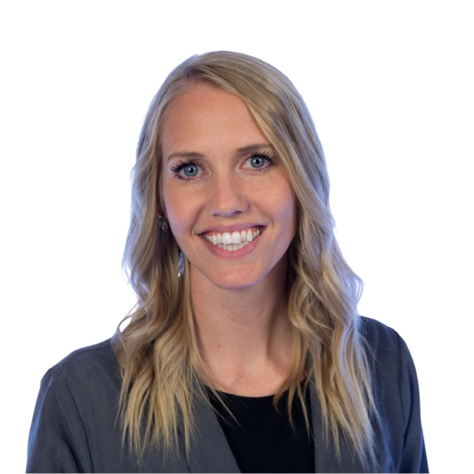 A woman with long blonde hair wearing a gray blazer over a black top, smiling in front of a plain white background.