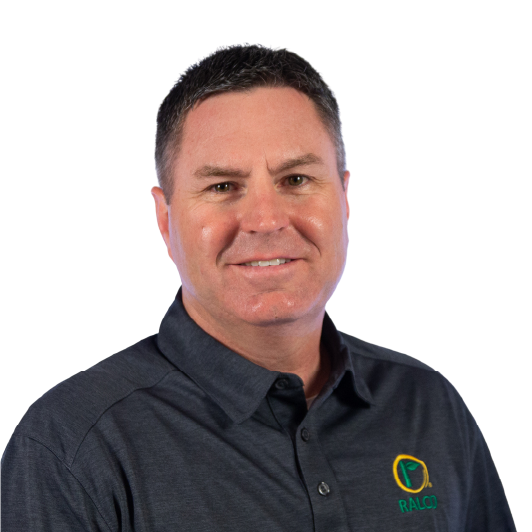 A man with short dark hair smiles at the camera. He is wearing a dark gray collared shirt with a green and yellow logo on the left side that reads "RALCO." The background is plain white.
