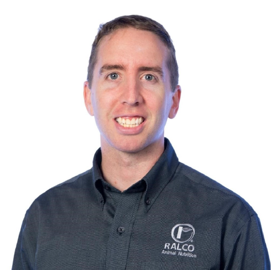 A man with short light brown hair smiles at the camera, wearing a dark grey button-up shirt with the "RALCO Animal Nutrition" logo on it, against a white background.