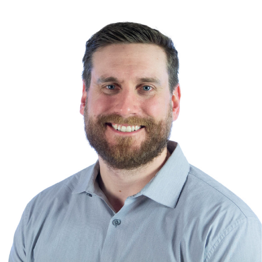 A man with light skin, brown hair, and a full beard smiles at the camera. He is wearing a light blue collared shirt and is photographed against a plain white background.