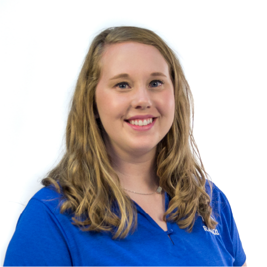 A woman with long, wavy, light brown hair smiles at the camera. She is wearing a blue collared shirt and a necklace, standing in front of a plain white background.