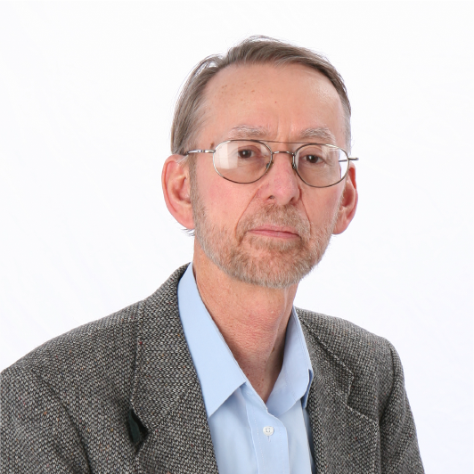 A middle-aged man with short gray hair, a beard, and glasses, wearing a gray tweed jacket over a light blue collared shirt, poses against a plain white background.