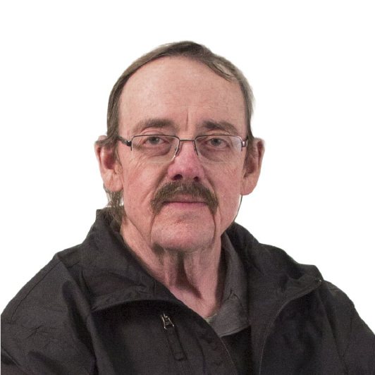 A middle-aged man with glasses, a mustache, and thinning hair wears a black jacket and looks directly at the camera, set against a plain white background.