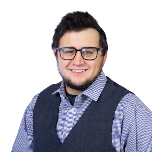 A smiling person with short dark hair, glasses, and a beard is wearing a blue checkered shirt and a dark vest, posed in front of a plain white background.