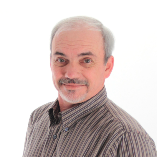 A middle-aged man with short gray hair and a trimmed beard smiles slightly, wearing a brown striped button-up shirt against a plain white background.