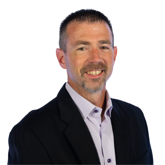 A man with short brown hair and a goatee, wearing a black blazer over a light purple collared shirt, smiles at the camera against a plain white background.