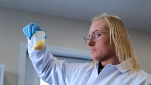 A scientist wearing safety glasses, blue gloves, and a white lab coat closely examines a clear plastic container with a yellowish substance inside.