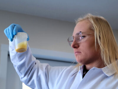 A scientist wearing safety glasses, blue gloves, and a white lab coat closely examines a clear plastic container with a yellowish substance inside.
