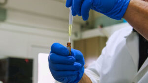 A person wearing blue gloves and a white lab coat uses a pipette to transfer liquid into a small glass vial in a laboratory setting.