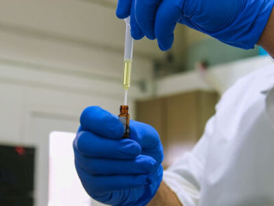A person wearing blue gloves and a white lab coat uses a pipette to transfer liquid into a small glass vial in a laboratory setting.
