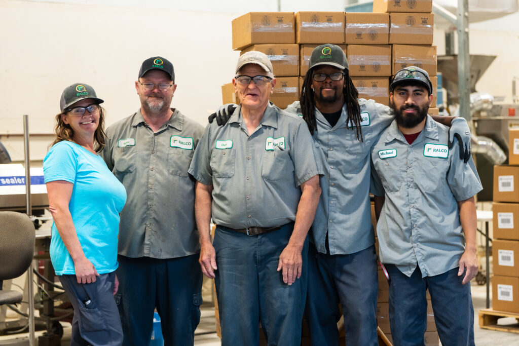 Five factory workers, four men in gray uniforms and one woman in a teal shirt, stand together smiling in a warehouse with stacked cardboard boxes in the background.