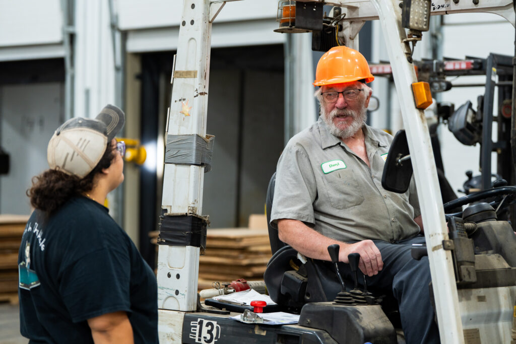 An older man wearing an orange hard hat and gray shirt sits on a forklift, talking to a woman in a black t-shirt and cap inside a warehouse with stacks of wood in the background.