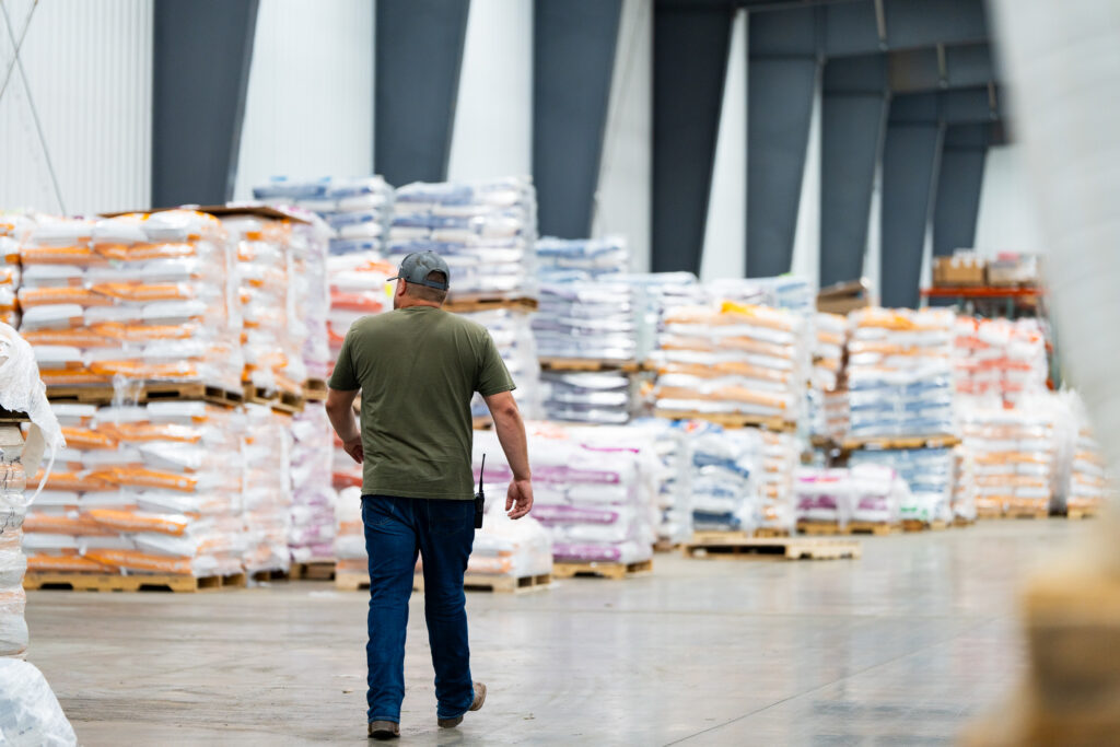 A man in a green shirt and cap walks through a warehouse with stacked pallets of packaged goods on both sides. The warehouse has high ceilings and concrete floors.