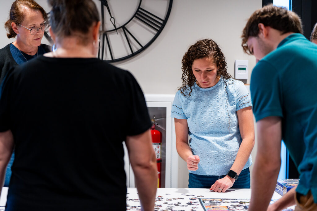 Four people are gathered around a table working on a jigsaw puzzle. A large wall clock and a fire extinguisher are visible in the background. The atmosphere appears focused and collaborative.