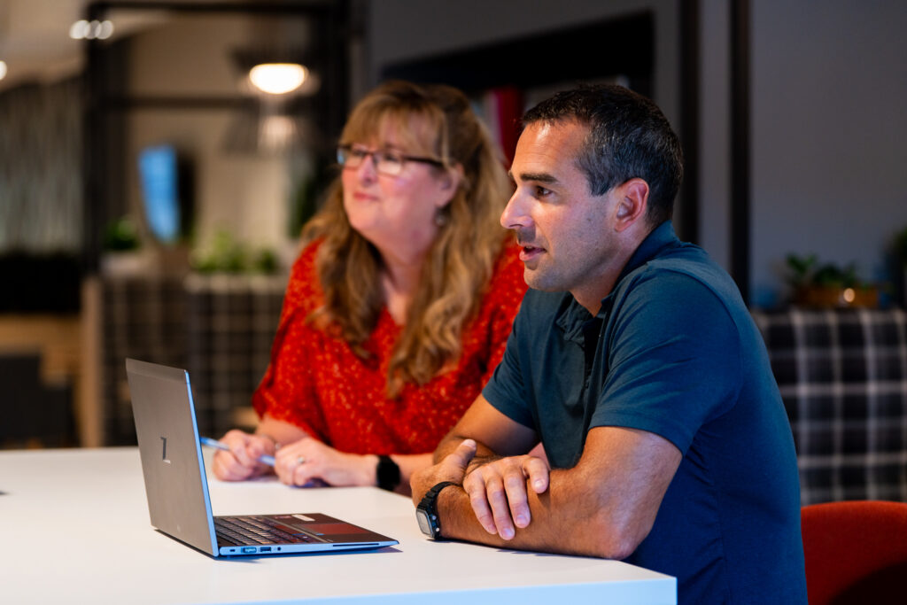 A man and a woman sit at a table, focused on a laptop. The woman has long blonde hair and wears glasses and a red blouse, while the man has short dark hair and wears a blue shirt. The setting appears to be an office.