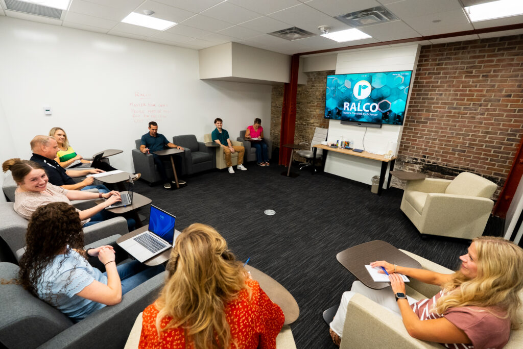 A group of people sit in a modern meeting room with armchairs arranged in a circle, facing a large screen displaying the RALCO logo. Some take notes on laptops, and a whiteboard is visible on the wall.
