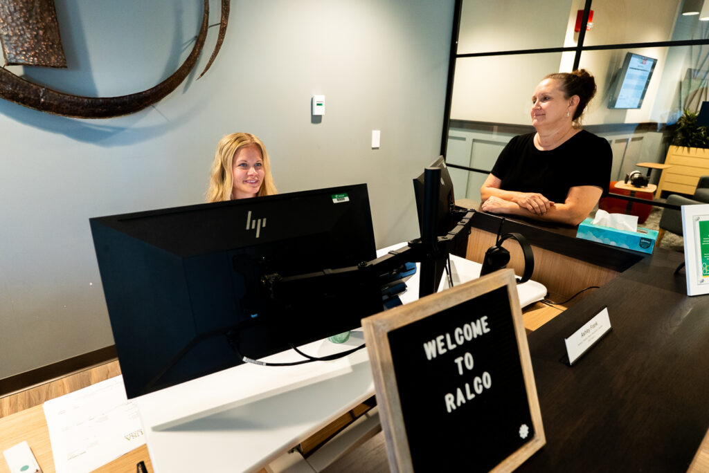 A woman sits at a reception desk smiling at another woman who is standing. A sign on the desk reads "Welcome to Ralco." The office environment is modern and well-lit.