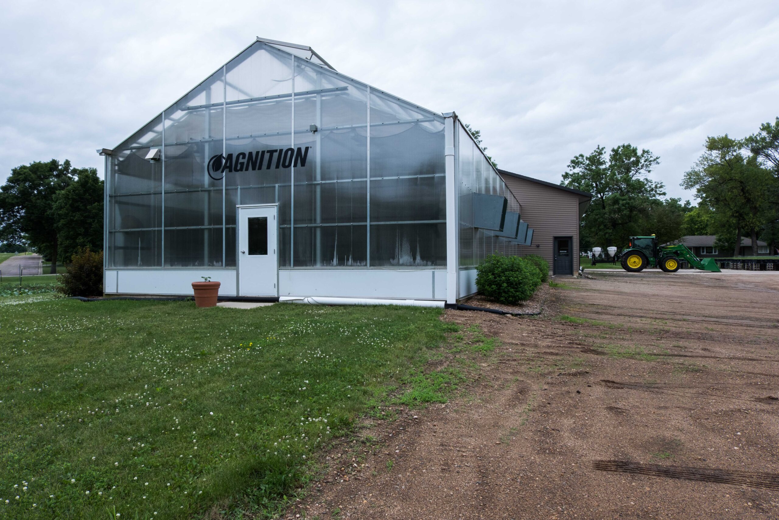 A glass greenhouse labeled "CAGnition" sits on grassy ground next to a dirt path, with a green tractor and trees visible in the background under a cloudy sky.