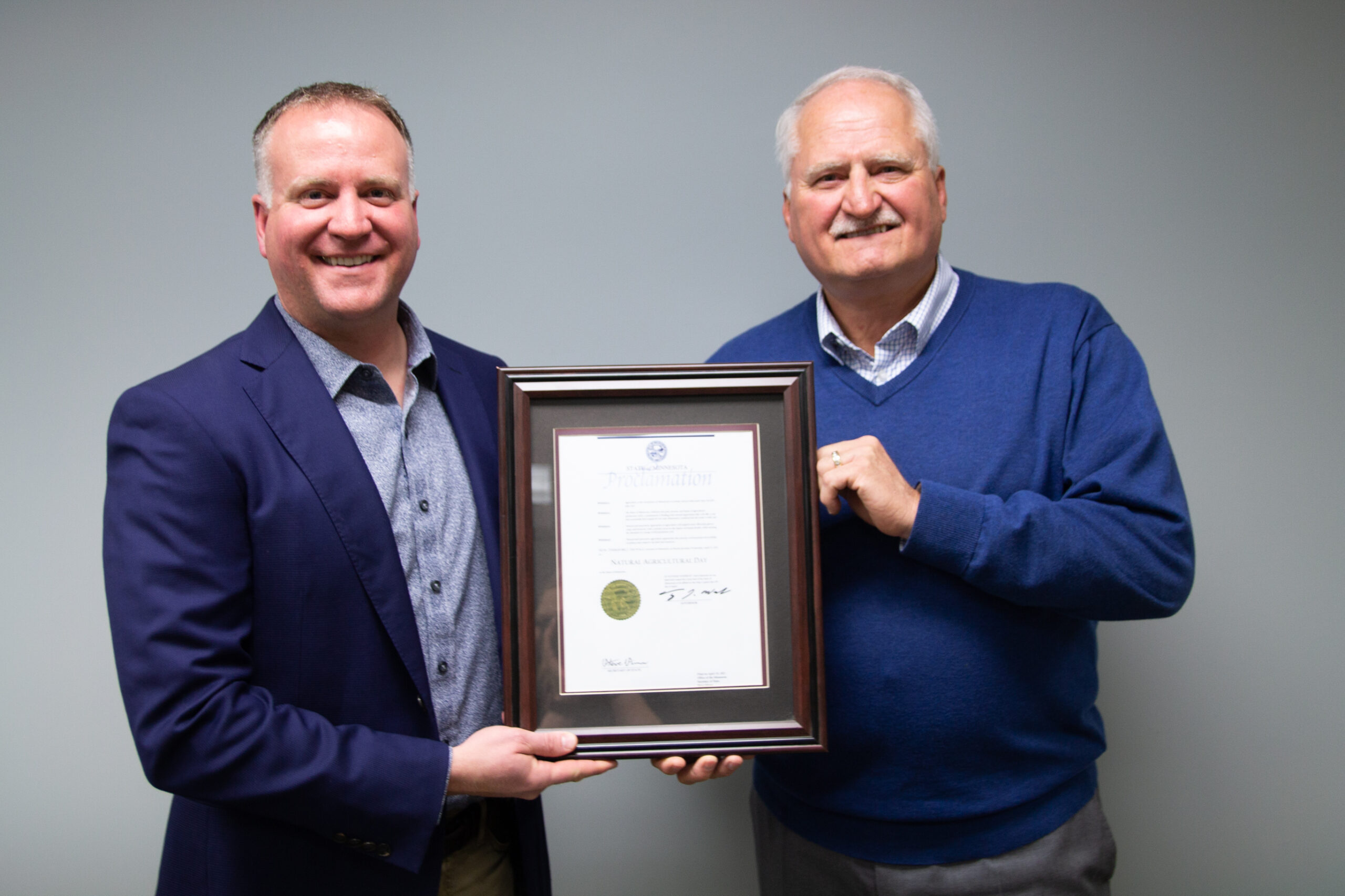 Two men in business attire stand side by side, smiling and holding a framed certificate against a plain gray background.