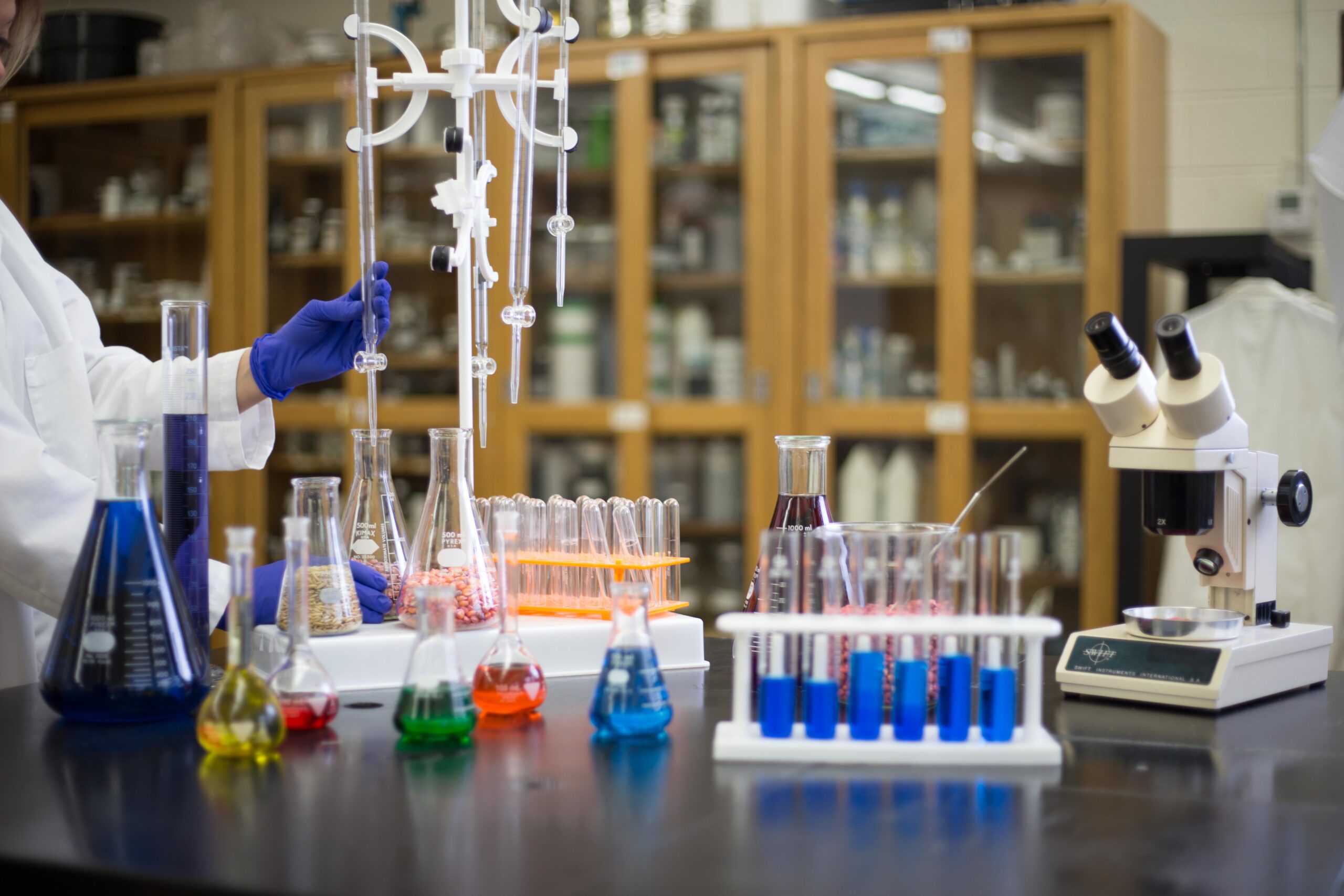 A scientist in a lab coat and blue gloves works with glassware and colorful liquids on a laboratory table, surrounded by test tubes, beakers, a microscope, and shelves filled with bottles and jars in the background.
