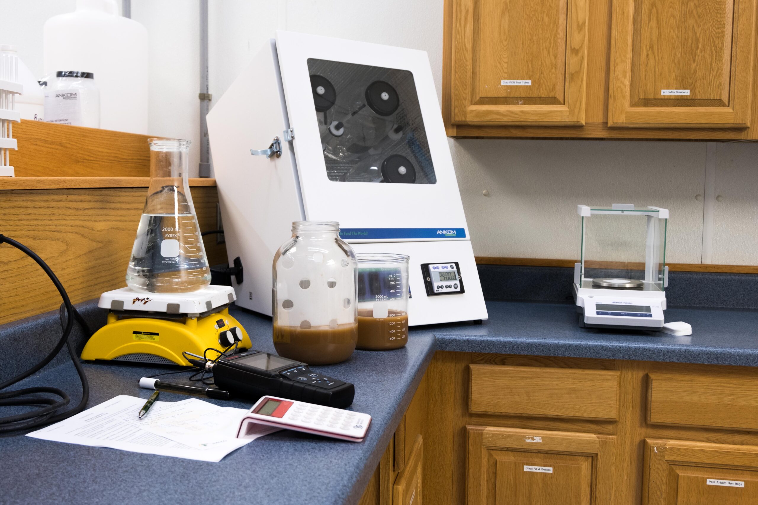 A laboratory counter with a beaker, glass jar of brown liquid, calculator, pH meter, stir plate, water incubator, and an electronic balance, surrounded by wooden cabinets and a sheet of paper.