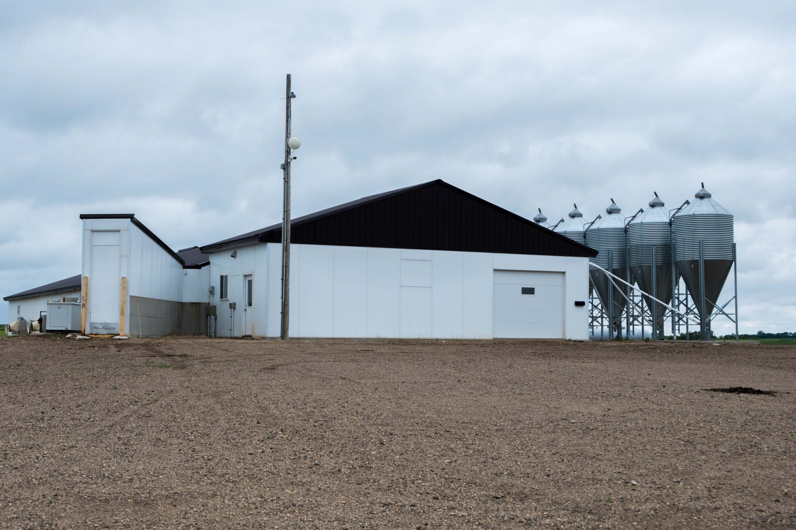 A large white barn with a dark roof stands on a gravel lot under a cloudy sky. Several tall metal silos are visible to the right of the barn. The scene is rural and open.