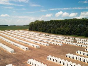 A large field contains many rows of small, white, plastic calf hutches, each separated evenly. In the background are green trees bordering the field under a partly cloudy blue sky.
